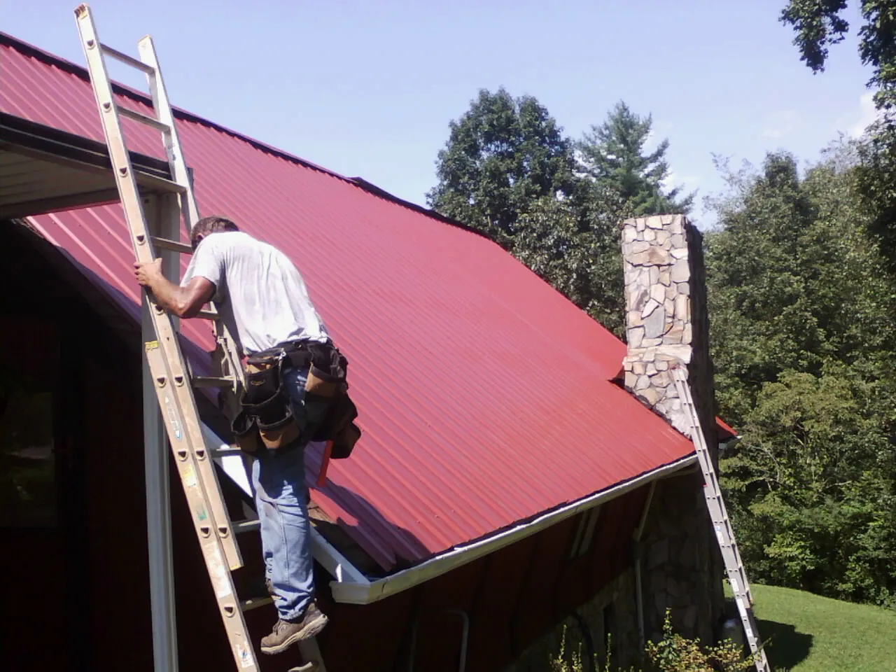 Roofer on a ladder beside a red metal roof and stone chimney — Soffit & Fascia Repair by Roofing Brothers in Mendham
